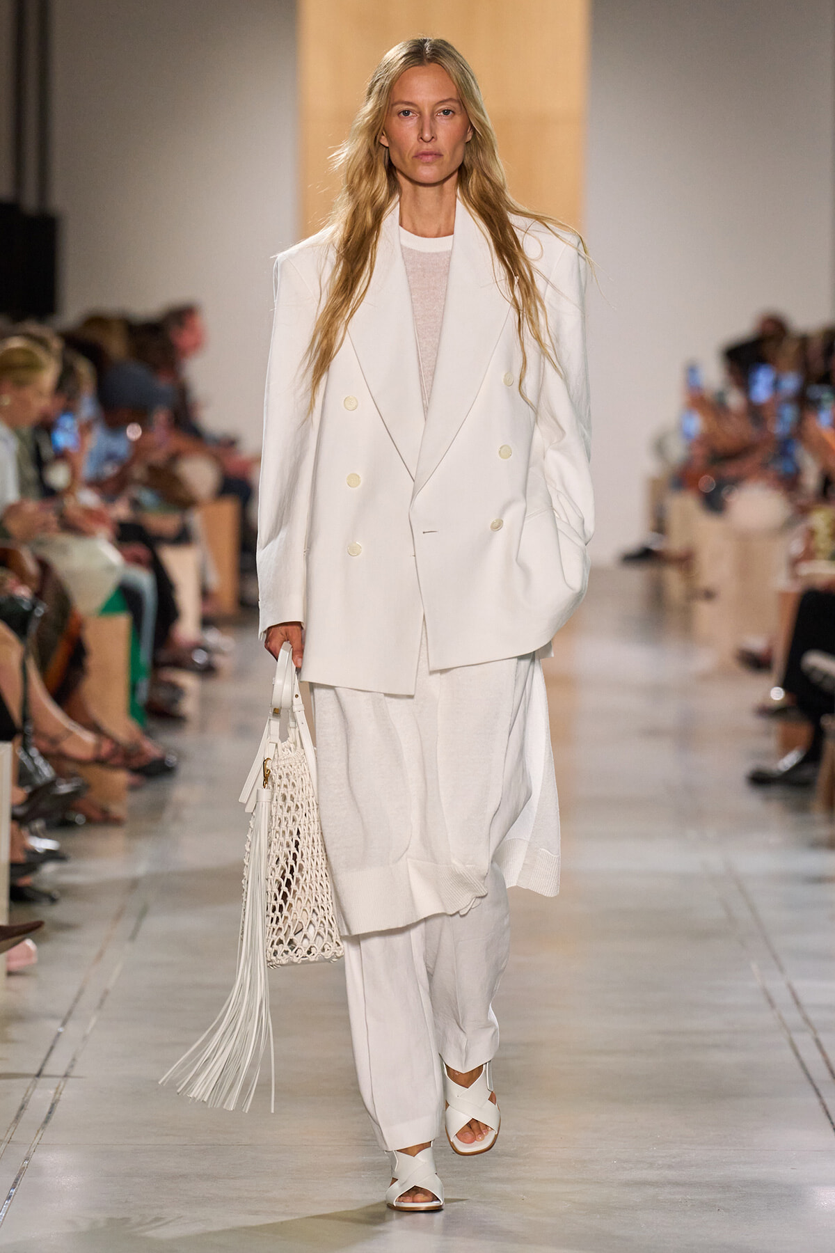 Model walks the runway in an oversized white double-breasted blazer and wide-leg white pants, carrying a white netted bag with fringe.