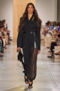 Model walks a runway in an all-black outfit: loose blazer with a wide belt and sheer black pants, carrying a black clutch.