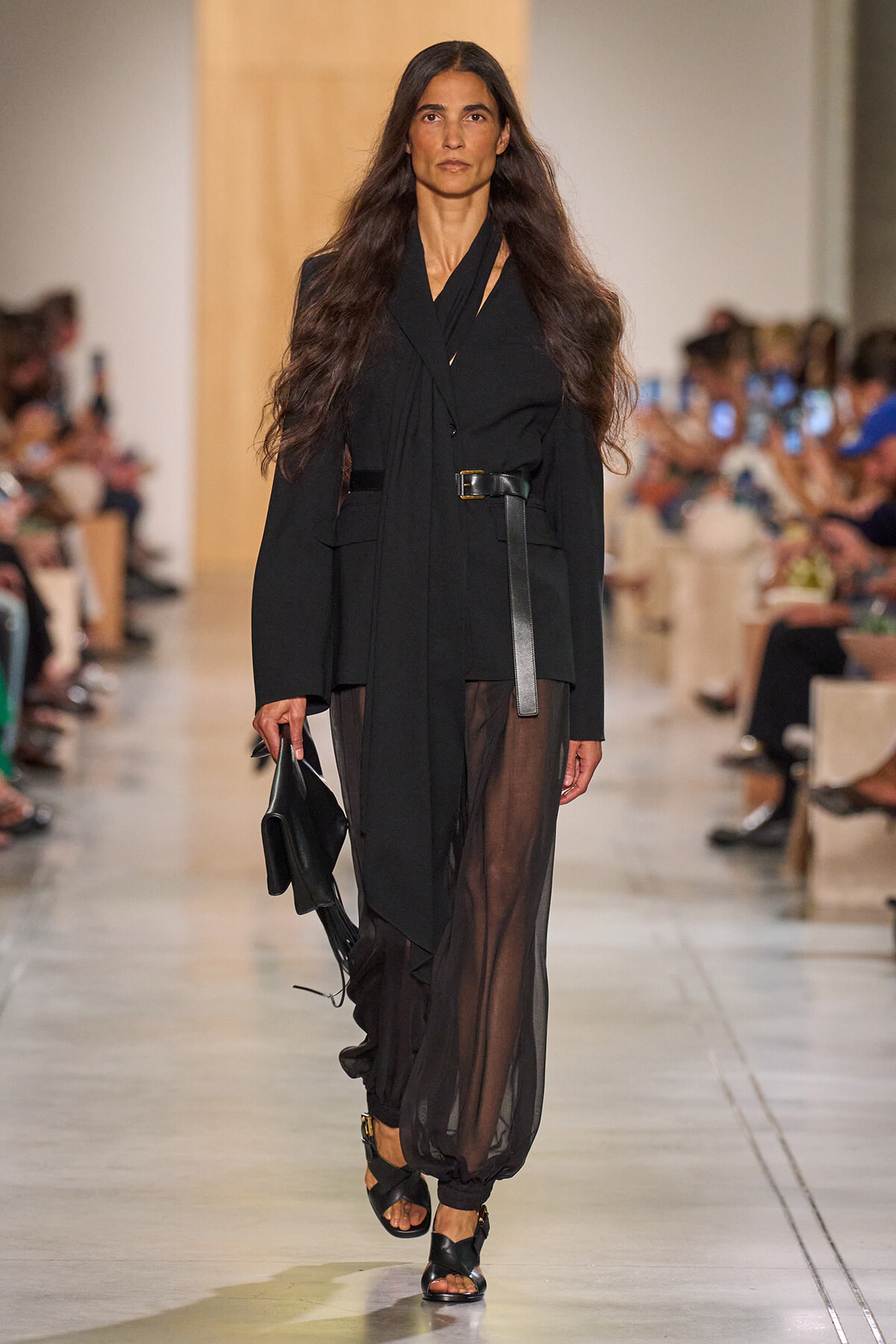 Model walks a runway in an all-black outfit: loose blazer with a wide belt and sheer black pants, carrying a black clutch.