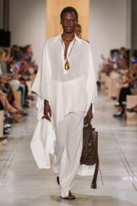 Model in a white flowing tunic and wide-leg pants walks a fashion show, accessorized with a large gold pendant necklace and a brown fringe bag on her side.