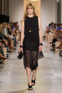 Model walking a fashion runway in a sleeveless black dress with a sheer, asymmetrical skirt and black sandals, accessorized with a long pendant necklace and chunky black wristbands.