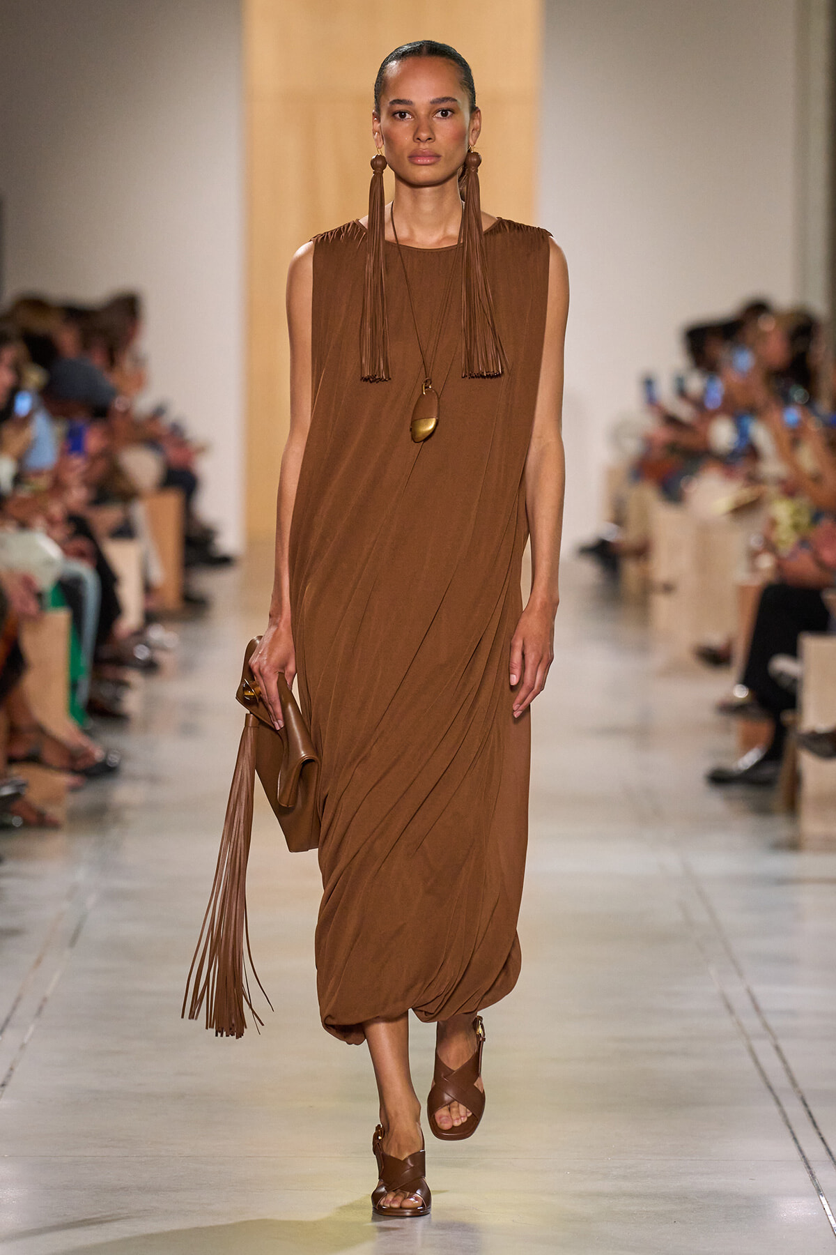 Model walks the runway in a brown sleeveless draped dress with long fringe earrings and a matching brown fringed bag and sandals.