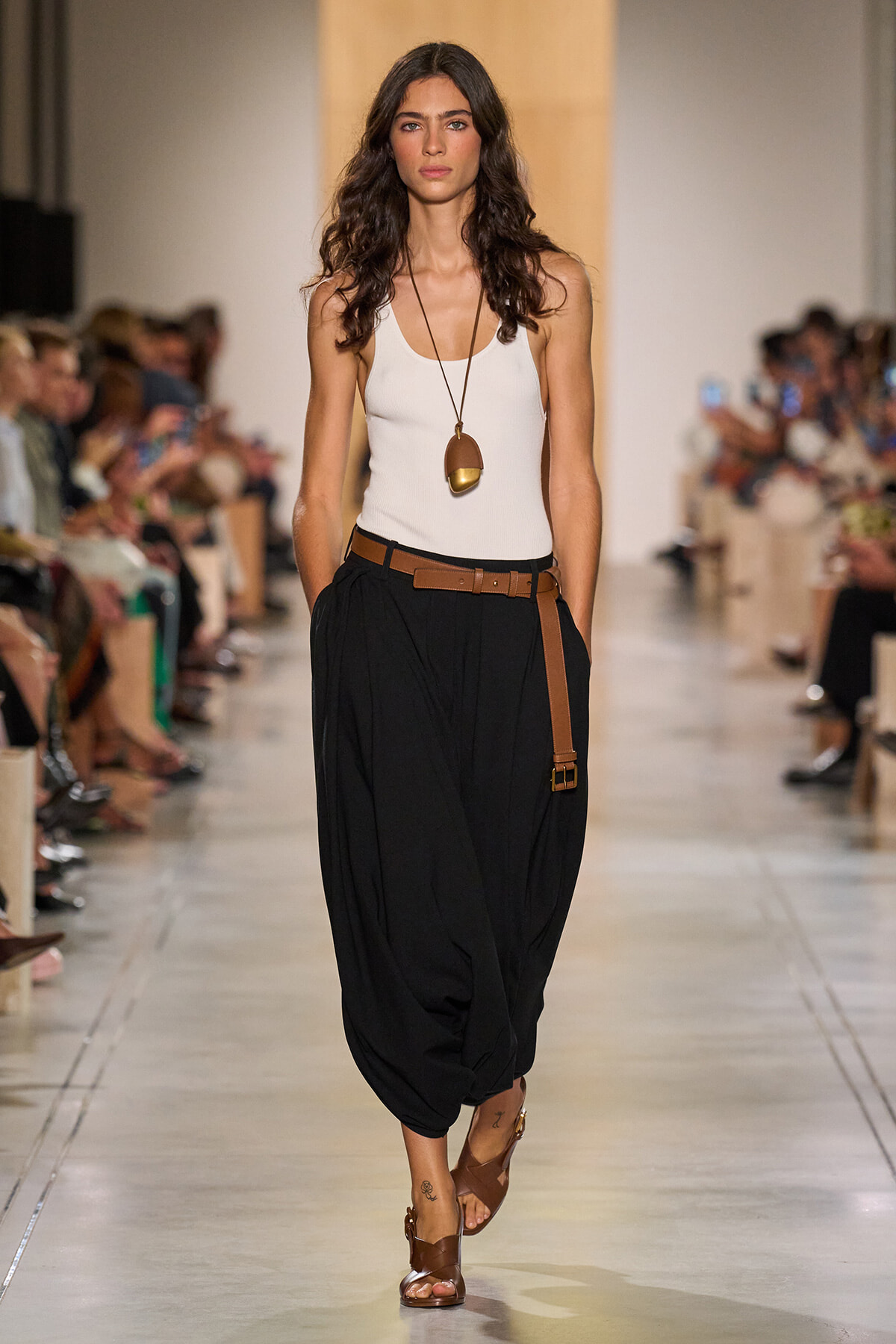 Model on the runway wearing a white tank top, black draped pants, a brown belt, brown sandals, and a long pendant necklace.