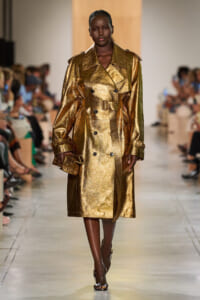 Model walks a runway wearing a gold metallic trench coat with a matching belt and dark skin tone, minimalist setting, blurred audience in background.