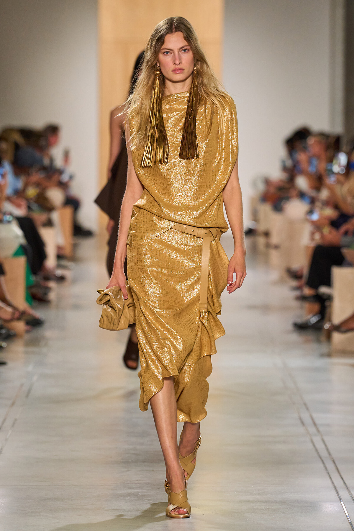 Model walking a gold metallic draped dress on a runway, holding a beige clutch and wearing long fringe earrings.