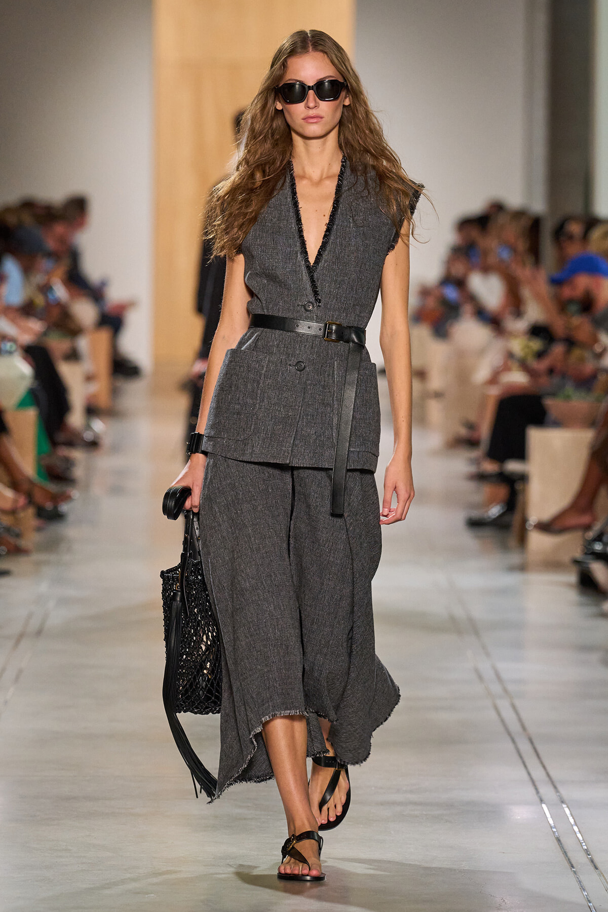 Model on a fashion runway wearing a gray textured sleeveless blazer with a matching wide-leg pant, black belt, and dark sunglasses, carrying a black net bag.