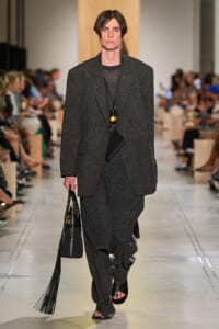 Male model on a runway wearing a charcoal textured suit, carrying a black tote with long fringe tassels and a layered pendant necklace visible.