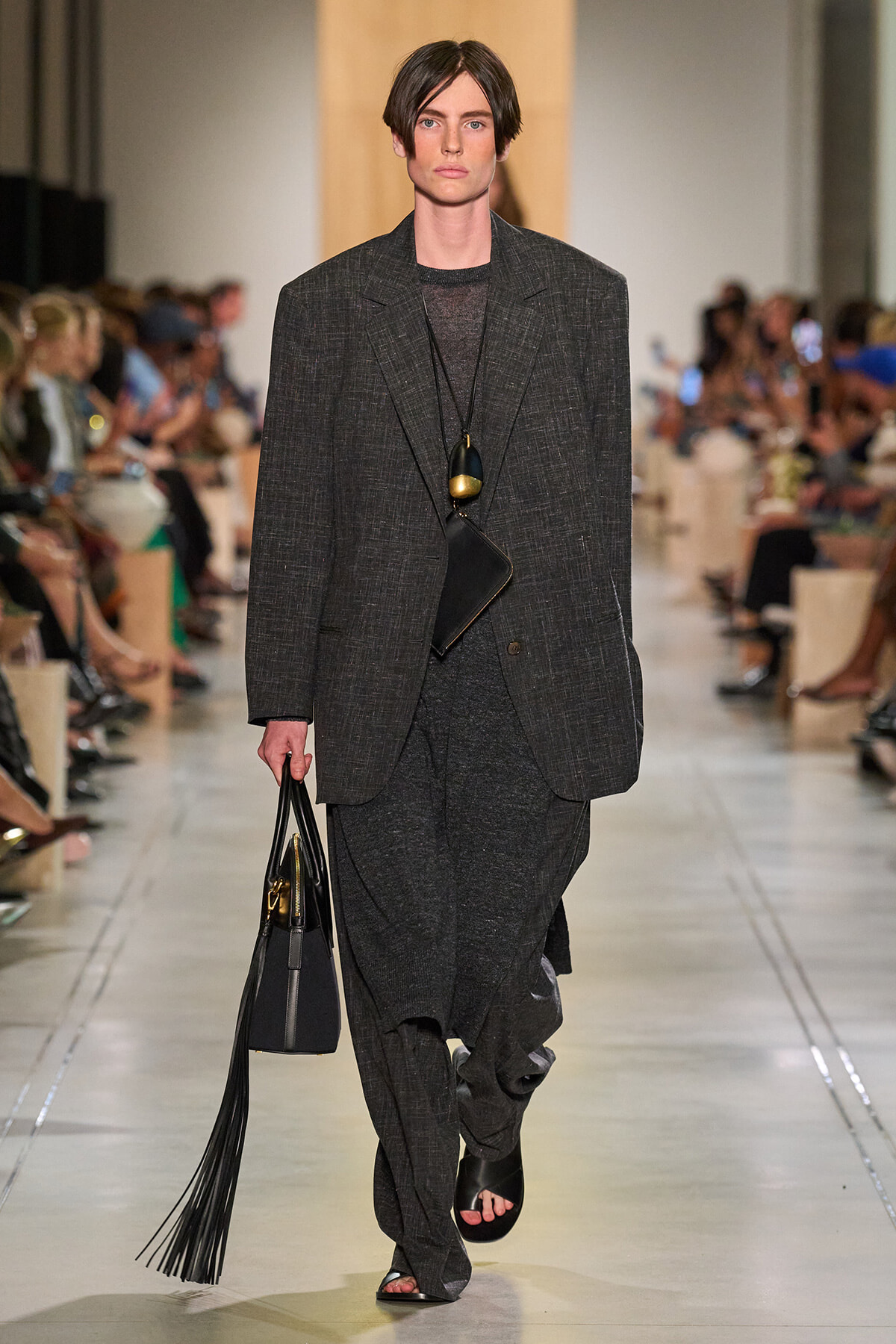 Male model on a runway wearing a charcoal textured suit, carrying a black tote with long fringe tassels and a layered pendant necklace visible.