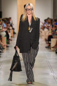 Runway model wearing an oversized charcoal blazer, striped wide-leg pants, black glasses, and a black netted handbag.