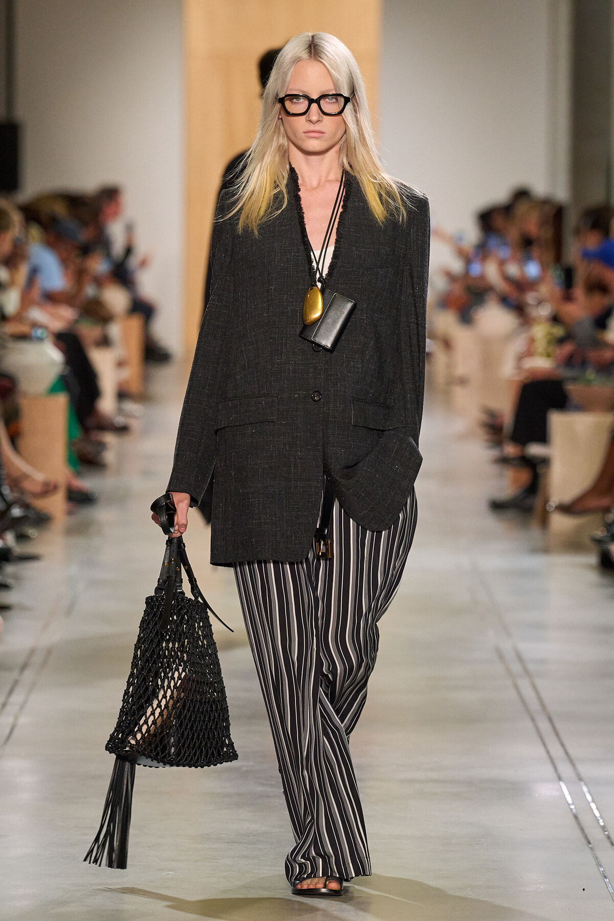 Runway model wearing an oversized charcoal blazer, striped wide-leg pants, black glasses, and a black netted handbag.