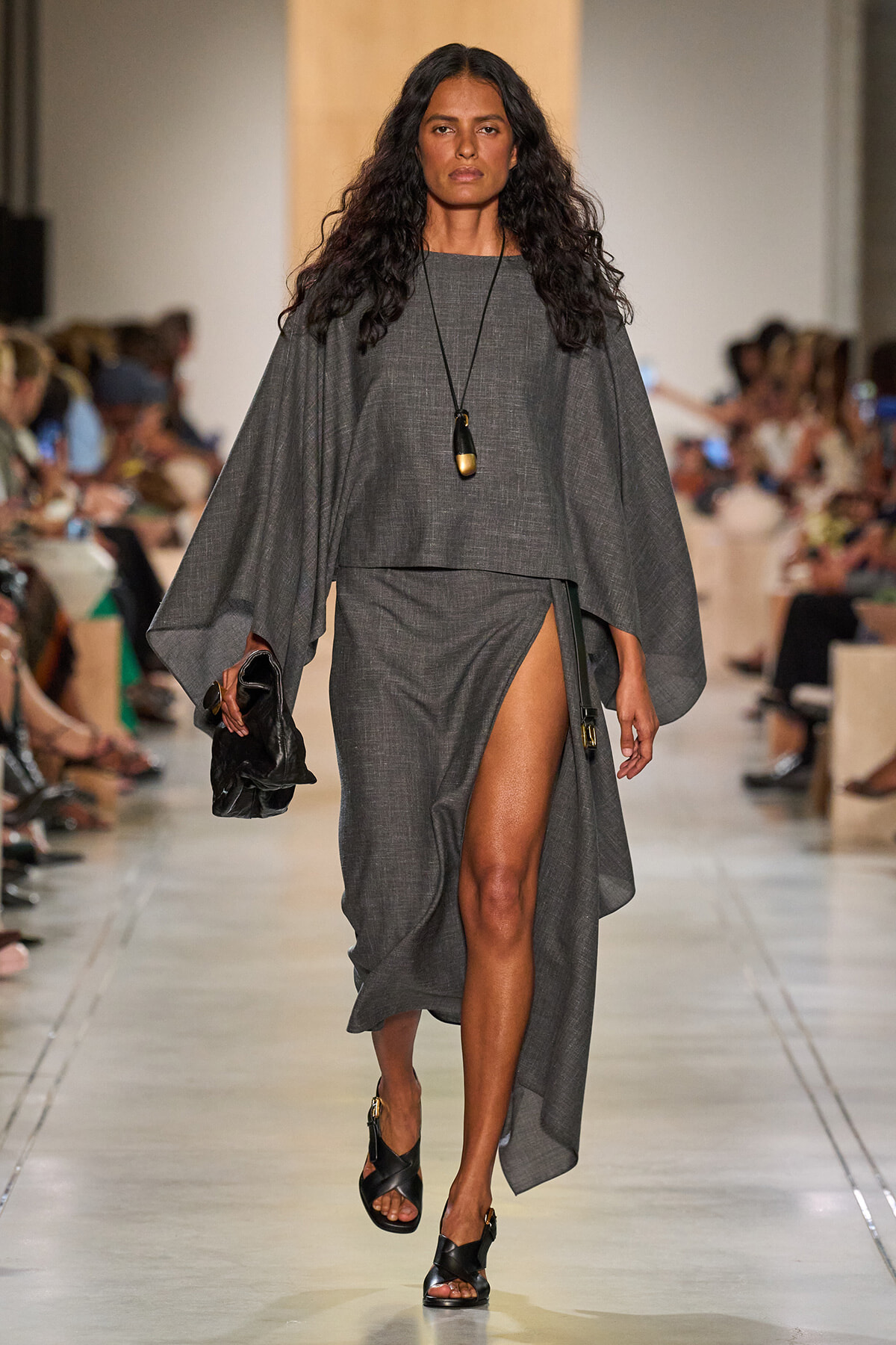 Model walks a runway in a gray cape-style top and matching wrap skirt with a high slit, black sandals, and a long pendant necklace.