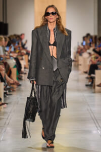 Model walks a runway in an oversized charcoal suit with striped shirt, black bra, and black sandals, carrying a textured black bag and wearing sunglasses.