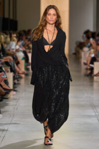 Model walking runway in a black wrapped top and sequined black skirt, barefoot sandals visible, audience in background.
