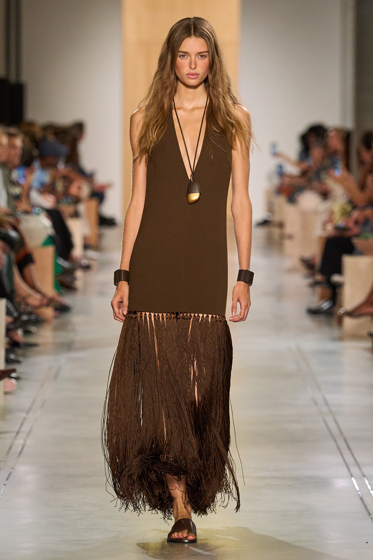 Model walks the runway in a brown sleeveless dress with a long fringe hem, paired with a large pendant necklace and black bracelets.