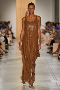 Model walks the runway in a bronze sequin sleeveless dress with a sheer brown hem, paired with tan sandals and chunky bracelets.