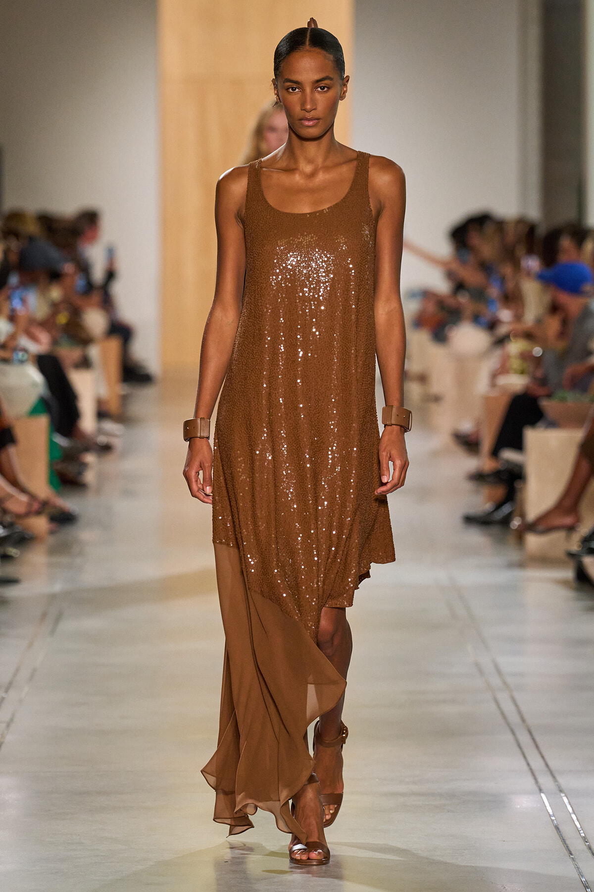 Model walks the runway in a bronze sequin sleeveless dress with a sheer brown hem, paired with tan sandals and chunky bracelets.