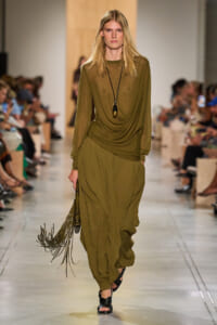 Model walking a runway in an olive draped ensemble: long-sleeve top and voluminous pants, with a black pendant necklace and fringe bag on her side.