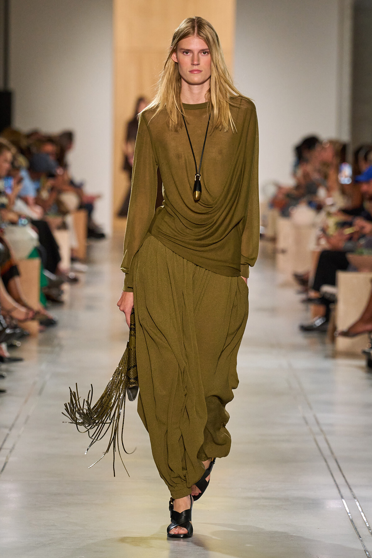 Model walking a runway in an olive draped ensemble: long-sleeve top and voluminous pants, with a black pendant necklace and fringe bag on her side.