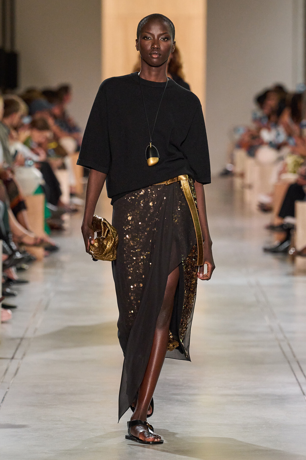 Model walking a runway in a black loose top and a gold-sequined wrap skirt with a metallic belt, holding a gold clutch and wearing flat sandals.