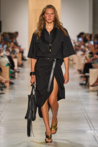 Model walks the runway in a black utility dress with a thigh-high slit, chunky black necklace, belt, and gold woven sandals, carrying a black woven bag.