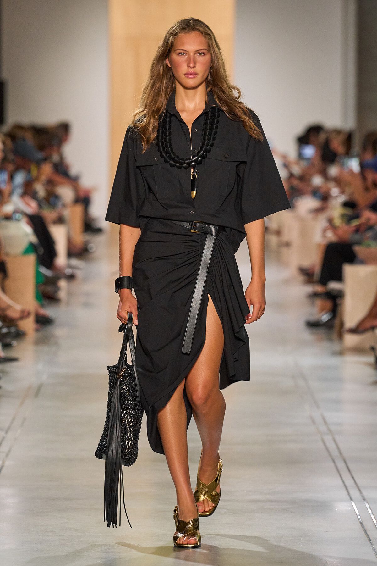 Model walks the runway in a black utility dress with a thigh-high slit, chunky black necklace, belt, and gold woven sandals, carrying a black woven bag.