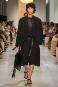 Male model walks down runway in oversized black trench, mesh top, and belt, carrying a fringed black bag and wearing sandals.