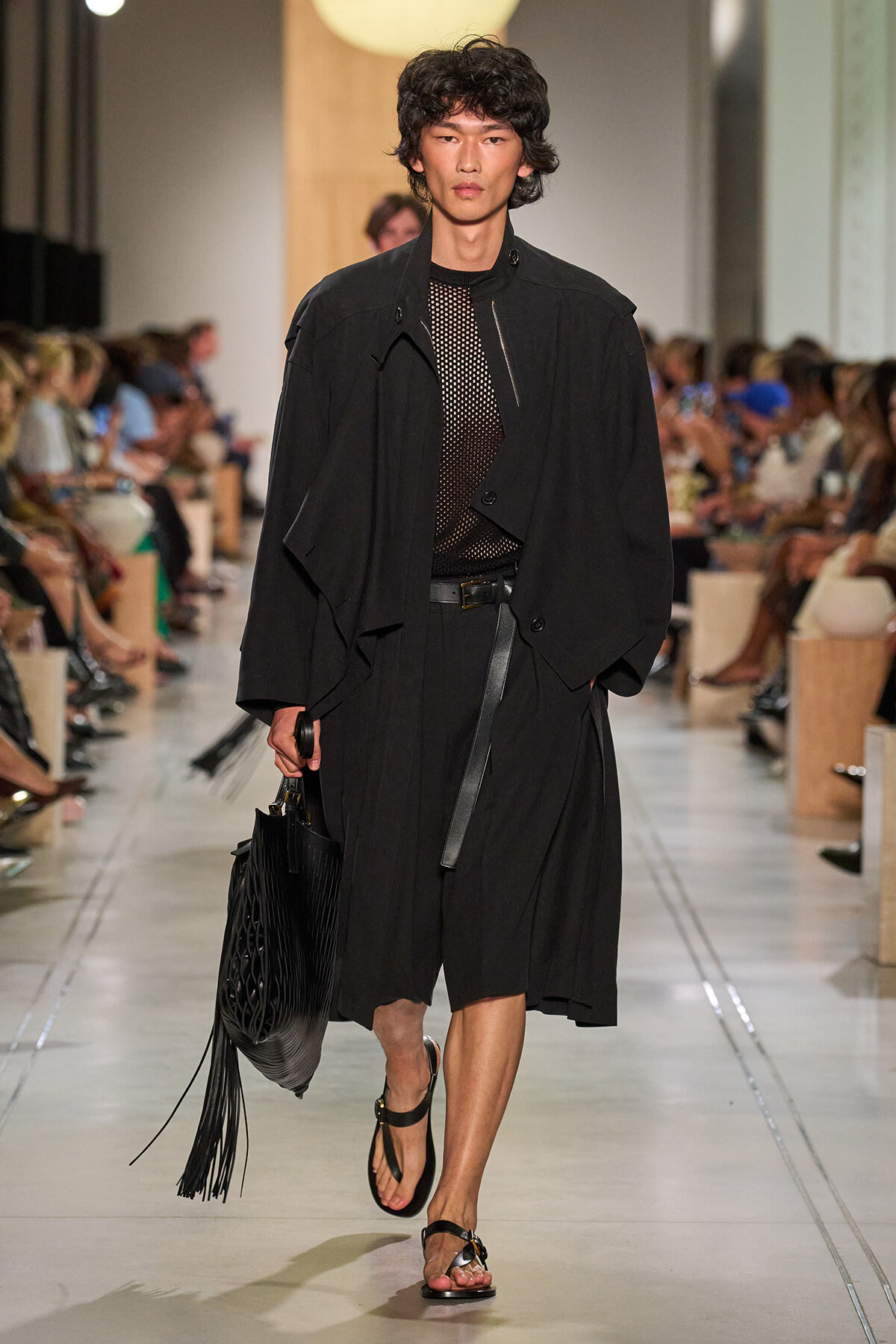 Male model walks down runway in oversized black trench, mesh top, and belt, carrying a fringed black bag and wearing sandals.