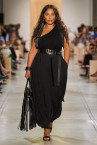 Model walks the runway in a black one-shoulder top, wide belt, and loose black trousers, carrying a fringed black handbag with long tassels.