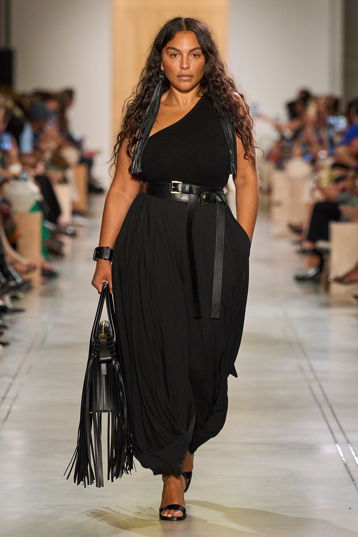 Model walks the runway in a black one-shoulder top, wide belt, and loose black trousers, carrying a fringed black handbag with long tassels.