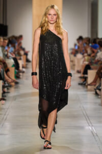 Model walking the runway in a black sequined one-shoulder dress with sandals, audience in the background.