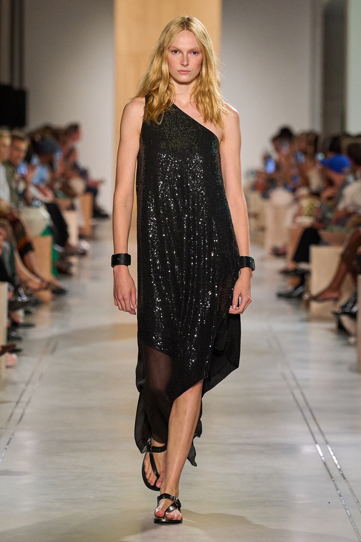 Model walking the runway in a black sequined one-shoulder dress with sandals, audience in the background.
