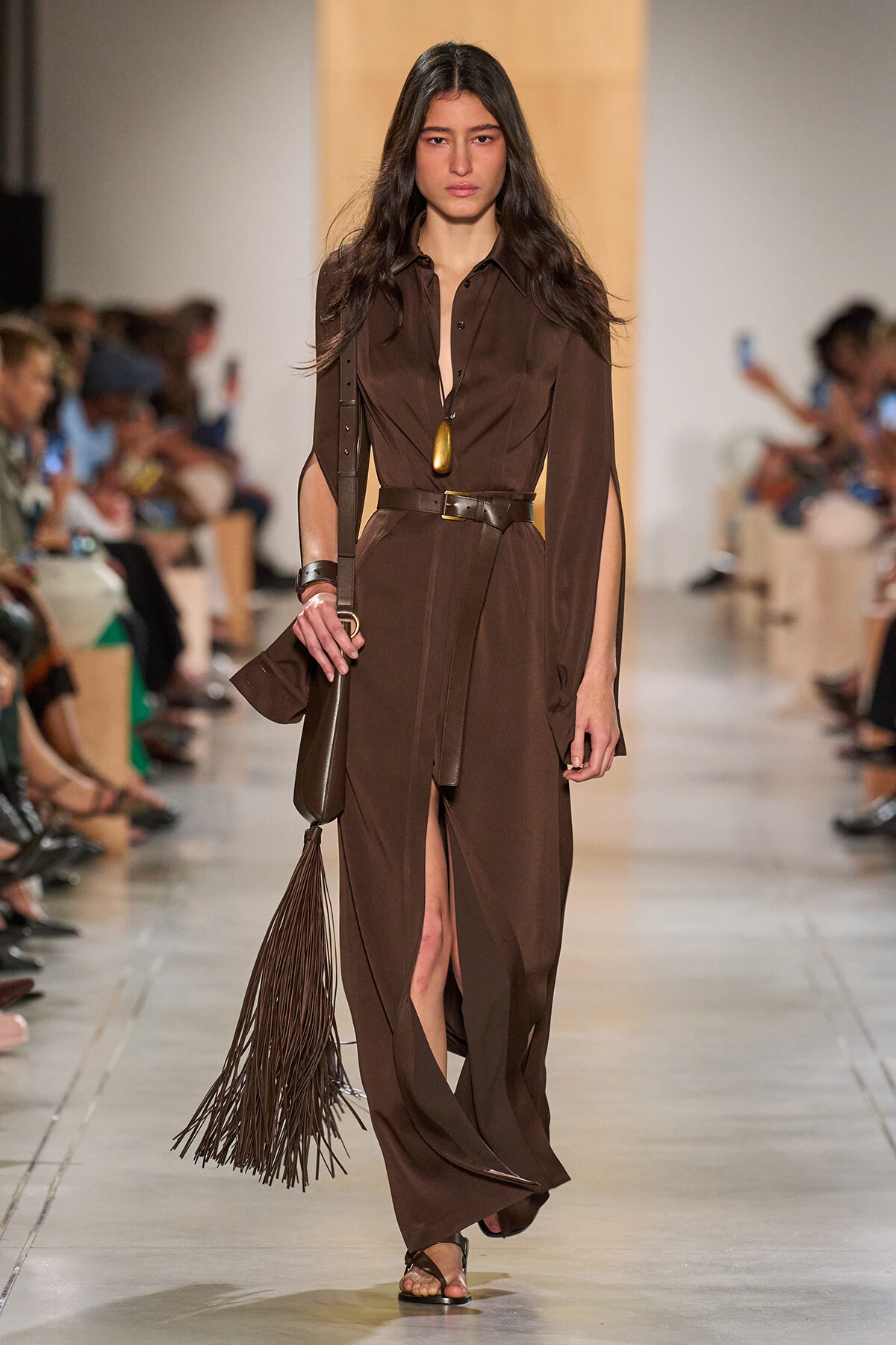 Model walks the runway in a brown, belted, long-sleeve dress with a front slit and fringe-bag accessories.