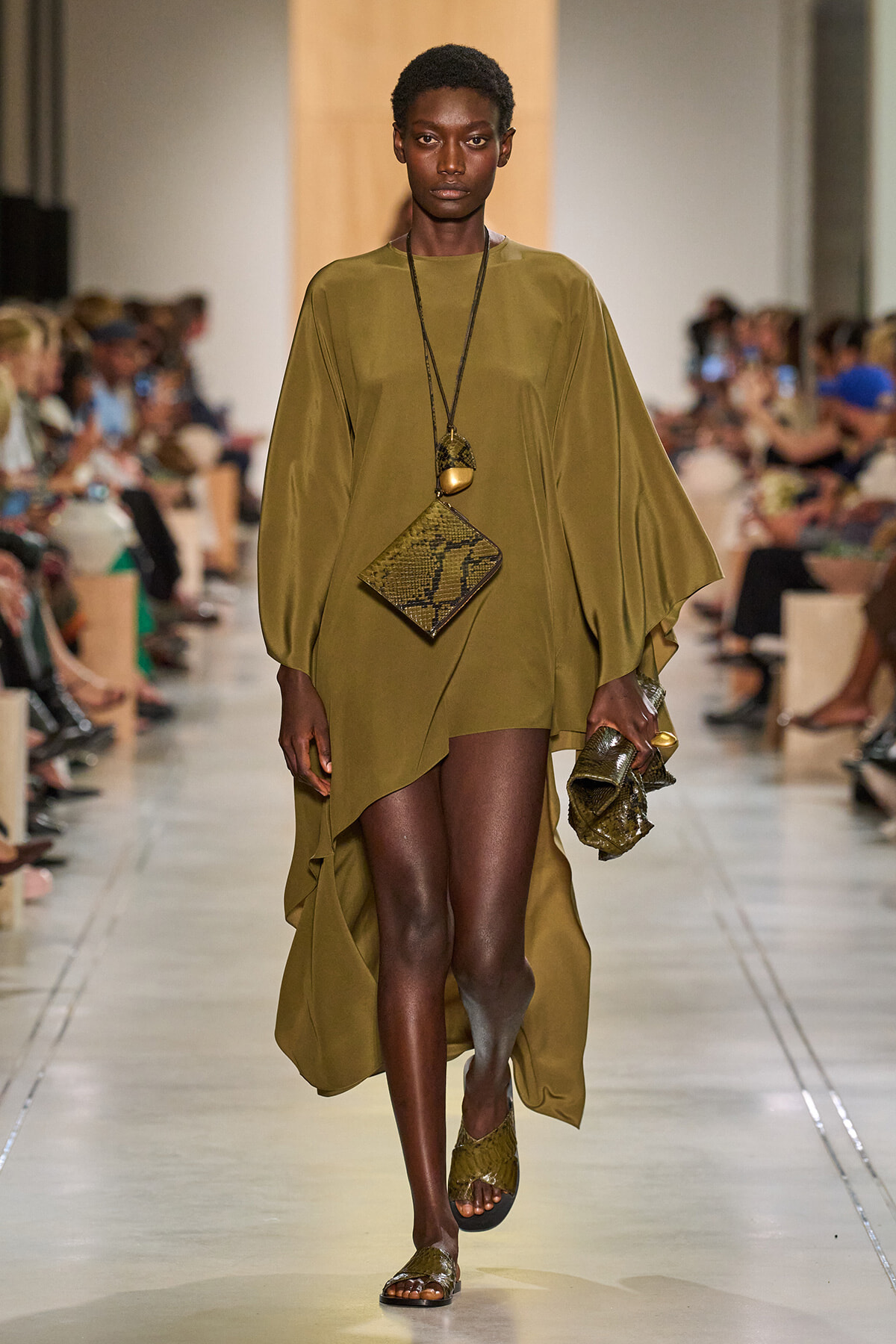 Model walking a runway in an olive green, draped dress with wide sleeves and matching snake-print accessories.