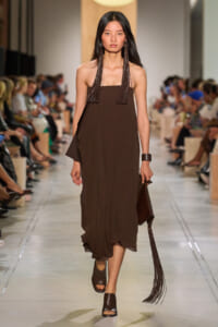 Model walking a runway in a brown strapless midi dress with long fringe earrings and a fringed clutch.
