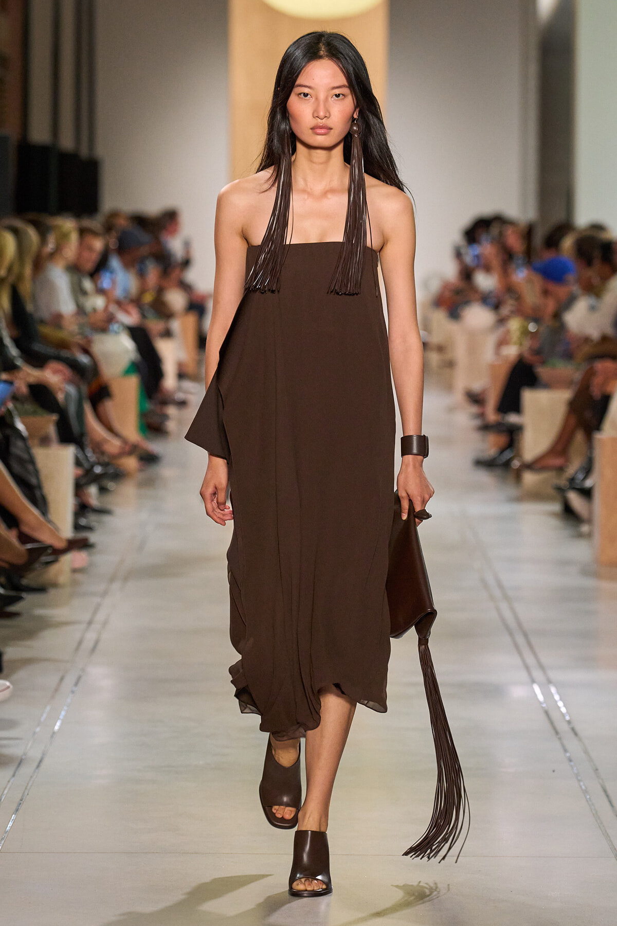 Model walking a runway in a brown strapless midi dress with long fringe earrings and a fringed clutch.