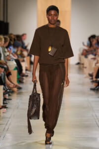 Model walking a runway in an all-brown outfit: oversized top, sheer brown pants, layered pendant necklace, and a net fringe bag.