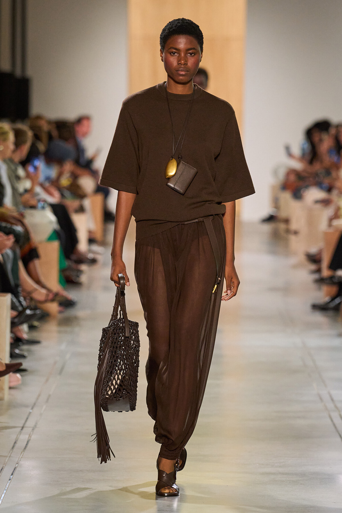 Model walking a runway in an all-brown outfit: oversized top, sheer brown pants, layered pendant necklace, and a net fringe bag.