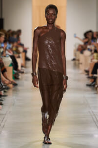 Model walks the runway in a one-shoulder brown sequined top and sheer brown pants, with a blurred audience in the background.