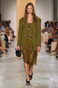 Model walks runway in an olive-green blazer and matching draped skirt, black sandals, and a long black necklace with a gold pendant, carrying a small black clutch.