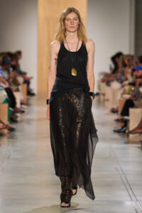 Model walking runway in black sleeveless top and shimmering black sequined pants with a sheer overlay, accessorized with a long pendant necklace and bracelets.