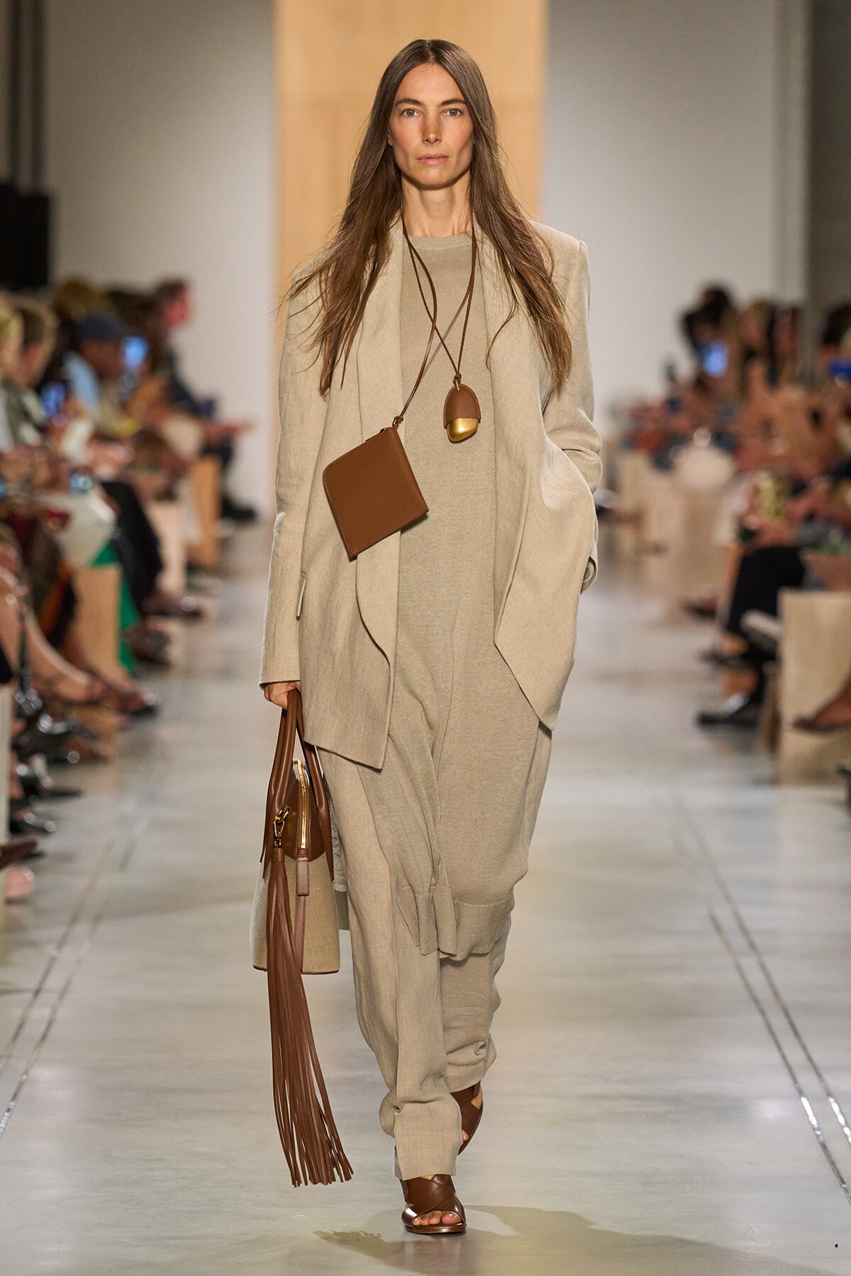 Model walks a runway in a monochrome beige ensemble with an oversized blazer and relaxed pants, accessorized with a brown fringe handbag and long pendant necklace.