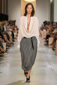 Model walking a runway in a white draped blouse, gray harem skirt, black belt bag, and black sandals at a fashion show event.