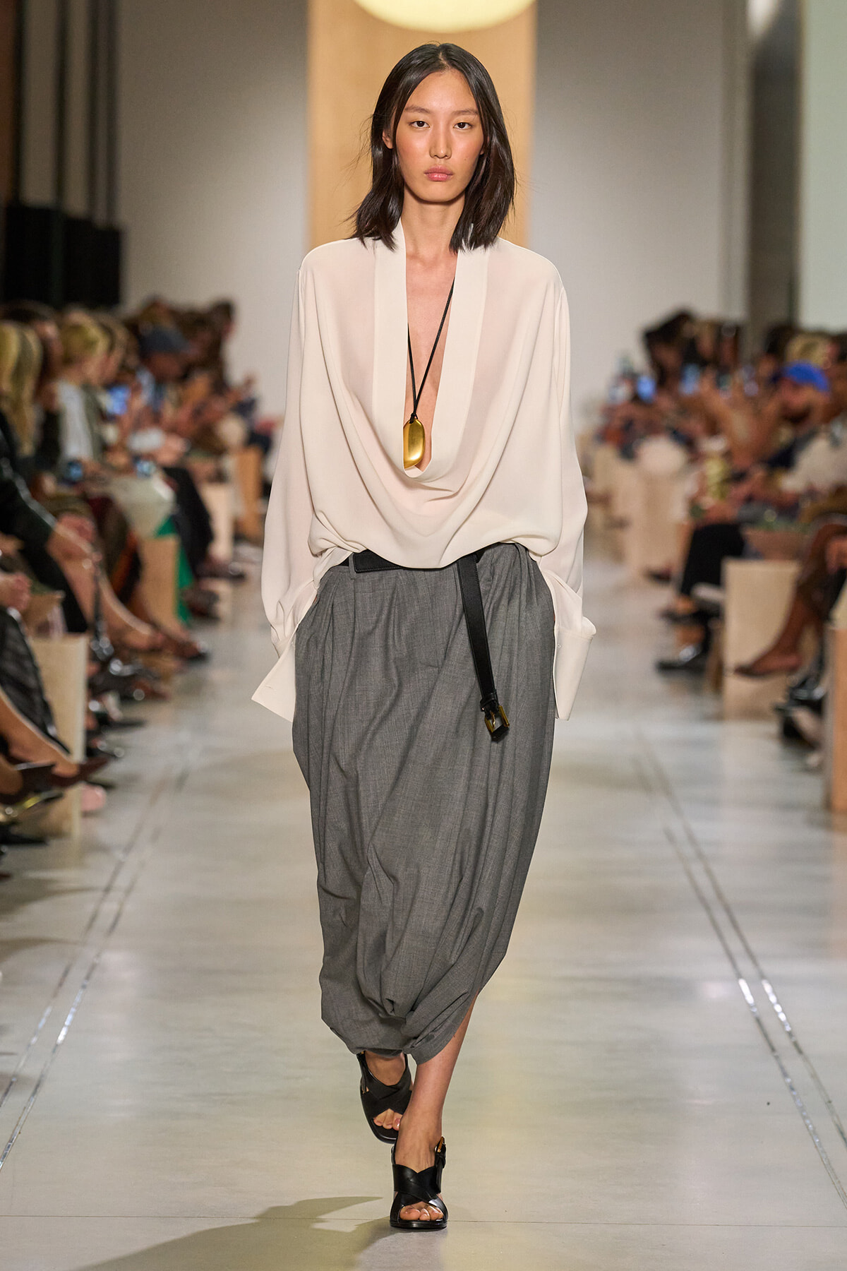 Model walking a runway in a white draped blouse, gray harem skirt, black belt bag, and black sandals at a fashion show event.