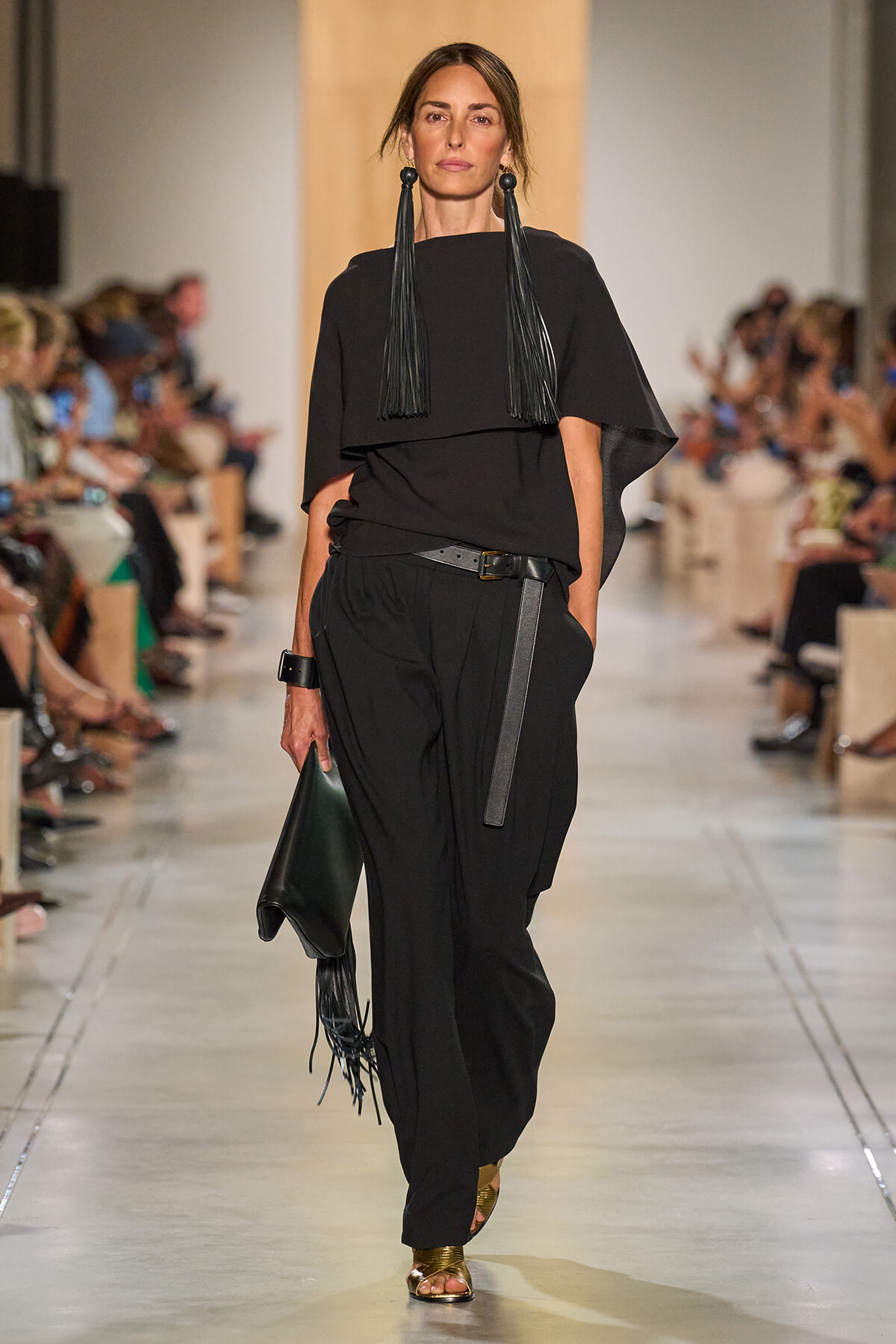 Model walking runway in a black, layered top and wide-leg pants with a belt and fringe earrings; holding a black clutch.