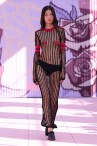 Model walking a runway in a black sheer fishnet outfit with red trim, black undergarments, and black heels against a pink mural backdrop.