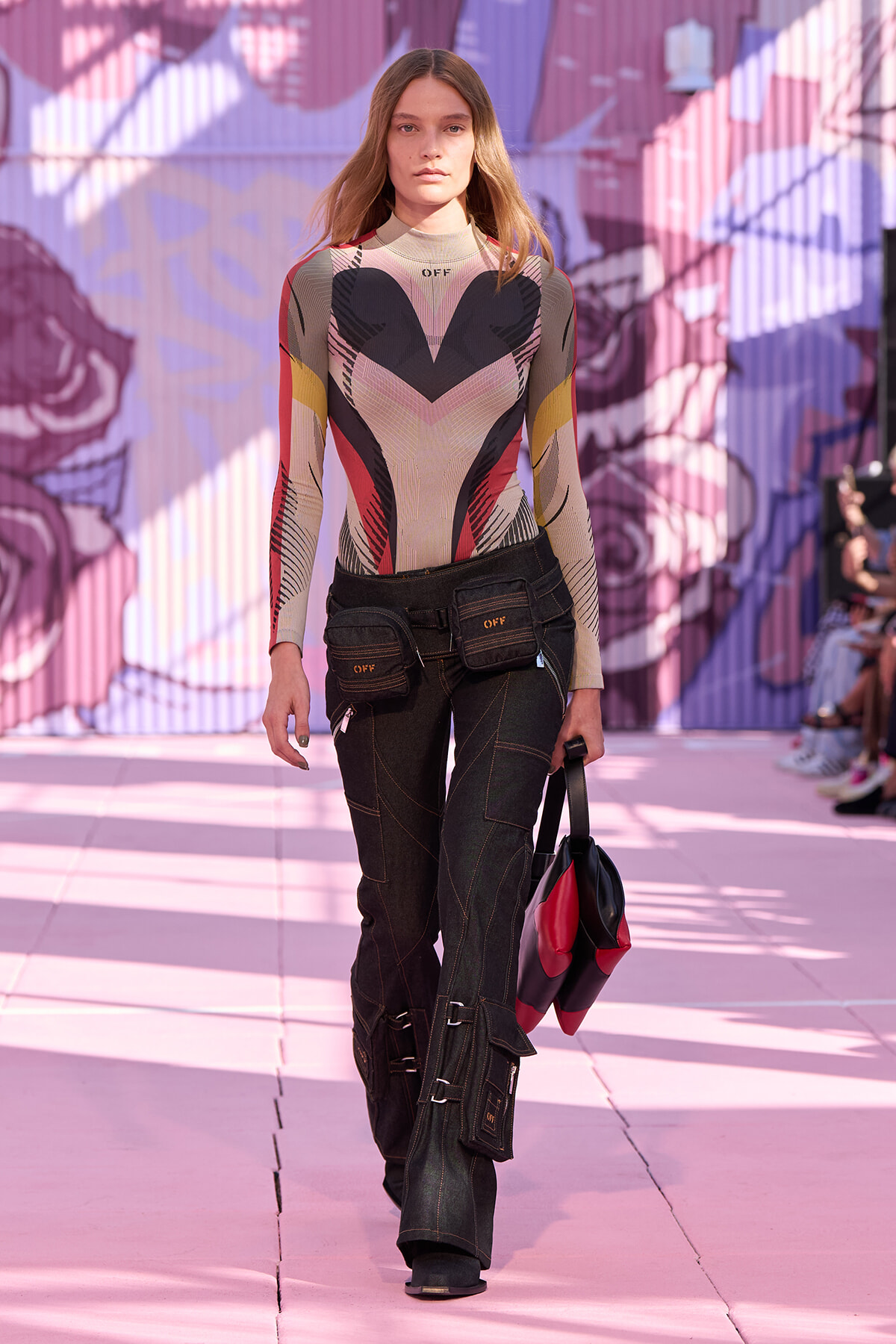 Model walking a fashion runway in a geometric-patterned bodysuit and dark cargo pants against a pink mural backdrop