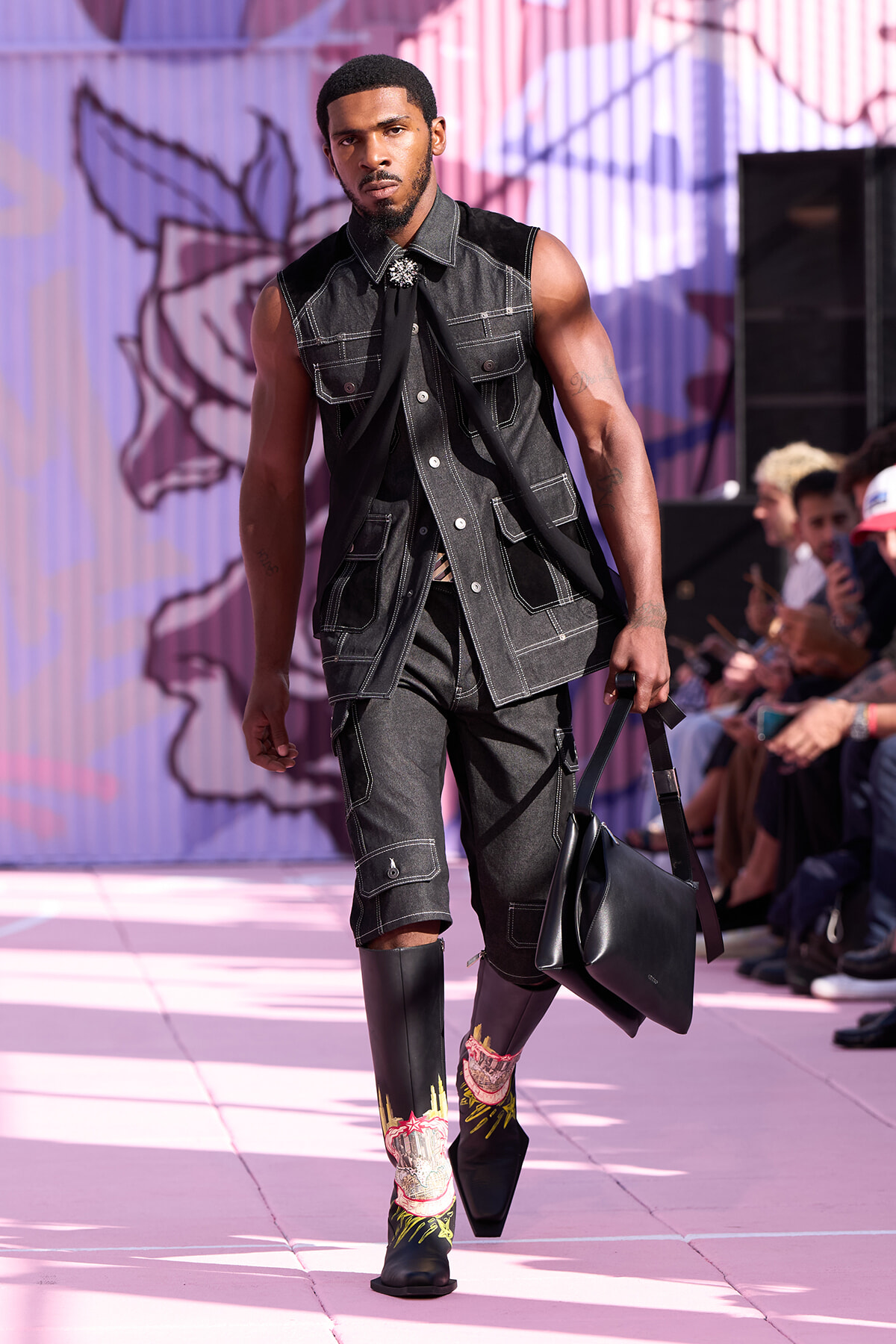 Male model walks a fashion show runway in a dark denim sleeveless vest and shorts with white stitching, carrying a black bag against a pink graffiti backdrop.