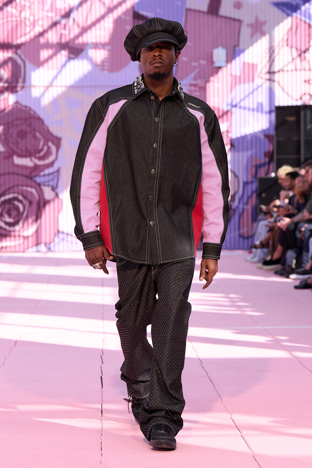 Male model walking a fashion runway in a black denim shirt with pink panels and studded pants, wearing a black cap under colorful backdrop.