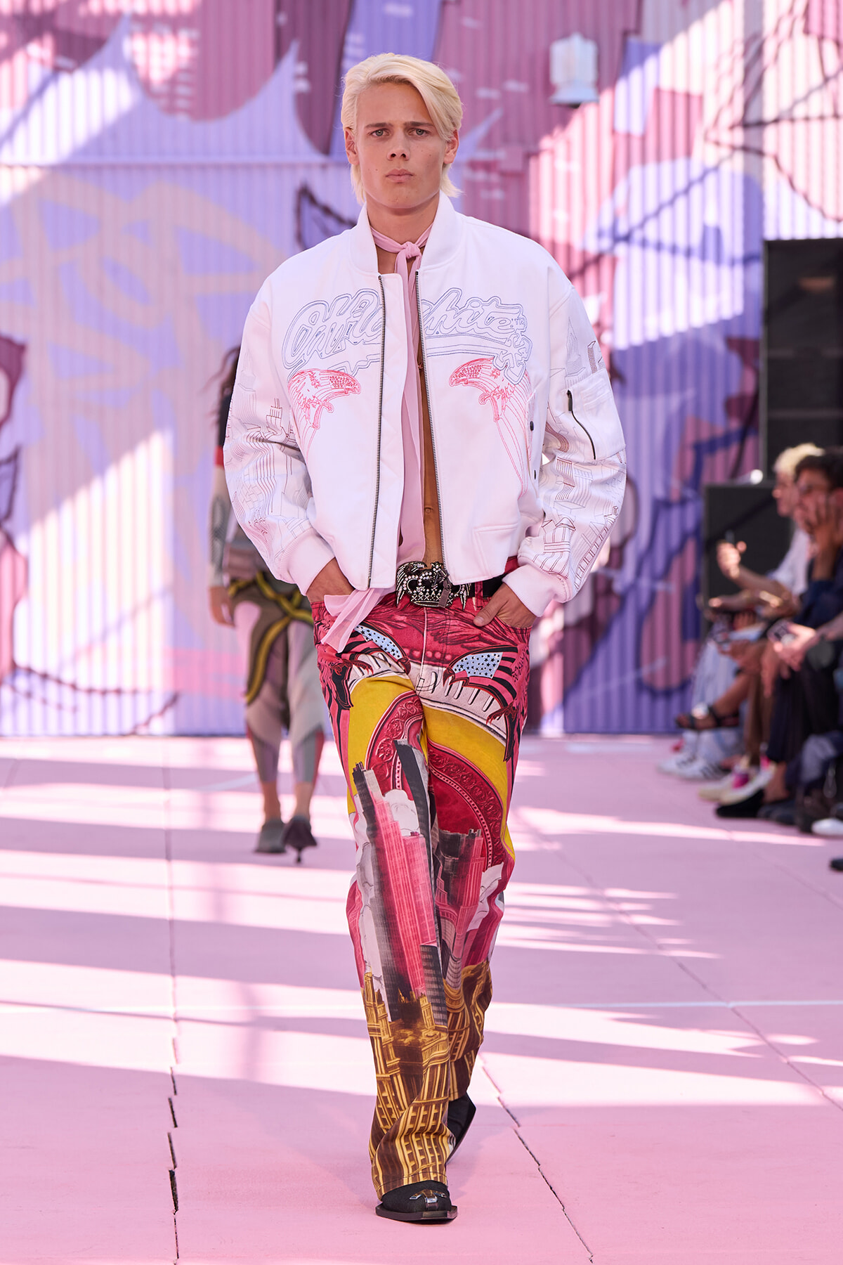 Model walking a runway in a pale pink bomber jacket and vibrant cityscape-print pants, pink-toned setting behind him.
