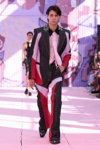 Male model wearing a color-block denim suit with pink shirt and scarf, walking on a pink runway backdrop.